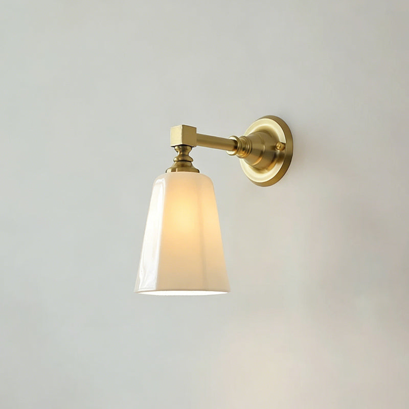 Close view of a gold Clérachic wall sconce lit, the tapered white glass shade glowing warm beneath a brass arm and round backplate.