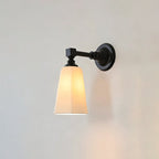 Black Clérachic wall sconce lit, with a tapered white glass shade glowing warm and casting a soft shadow on the pale wall.