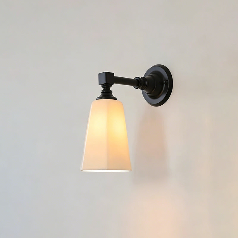 Black Clérachic wall sconce lit, with a tapered white glass shade glowing warm and casting a soft shadow on the pale wall.