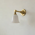 Gold Clérachic wall light with a flared bell-shaped white glass shade, switched off, on a pale grey wall.