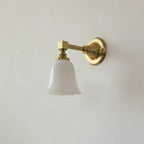 Gold Clérachic wall light with a flared bell-shaped white glass shade, switched off, on a pale grey wall.