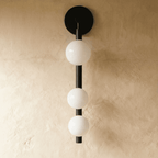 Clérachic black wall lamp with three opal glass spheres on a vertical rod, unlit, against a limewash wall.