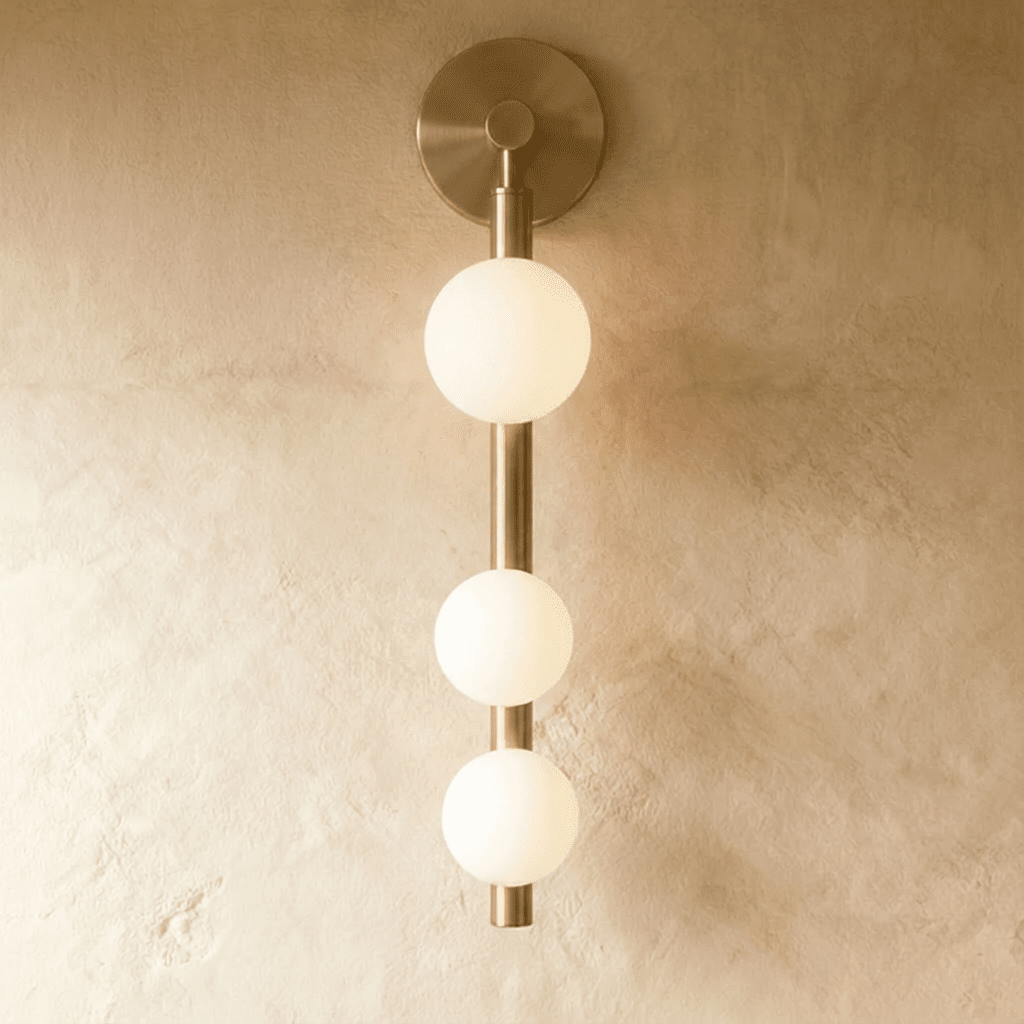 Clérachic wall lamp in satin brass with three glowing frosted glass spheres on a vertical rod and a round backplate.
