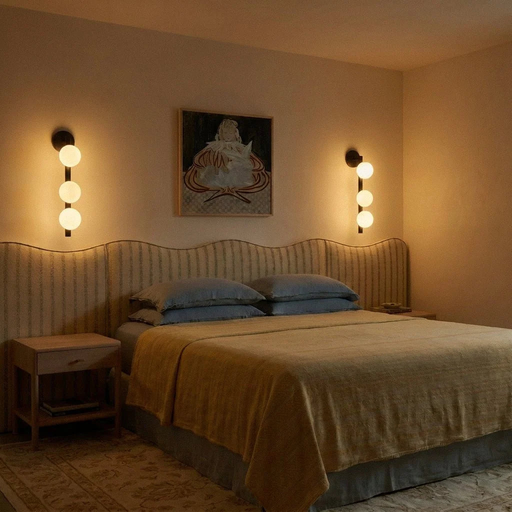 Pair of Clérachic black wall lamps lit warm, their frosted spheres glowing above a striped headboard in a dim bedroom.
