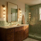 Clérachic brass wall lamps with glowing frosted spheres beside a brass-framed mirror above a walnut vanity and green tiles.