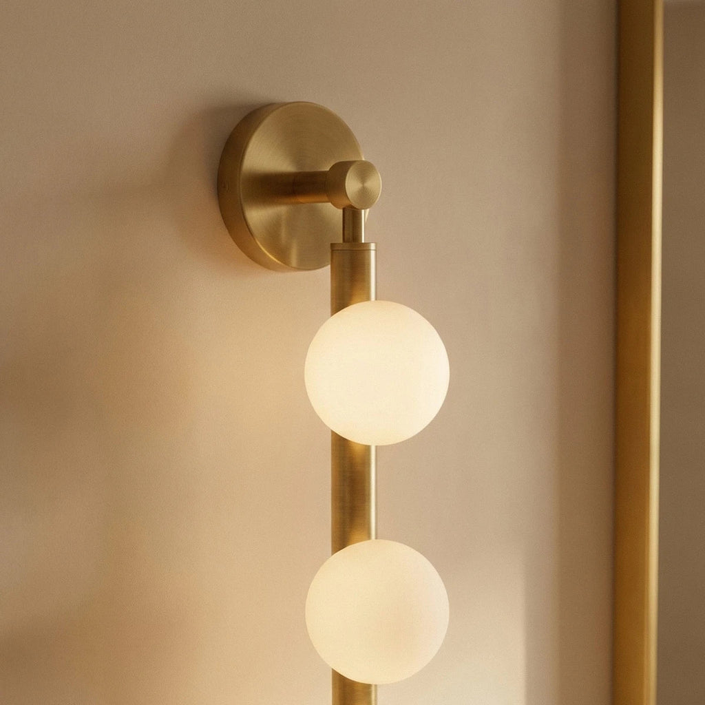 Clérachic brass wall lamp close up, a brushed brass backplate and rod holding two lit frosted glass spheres.