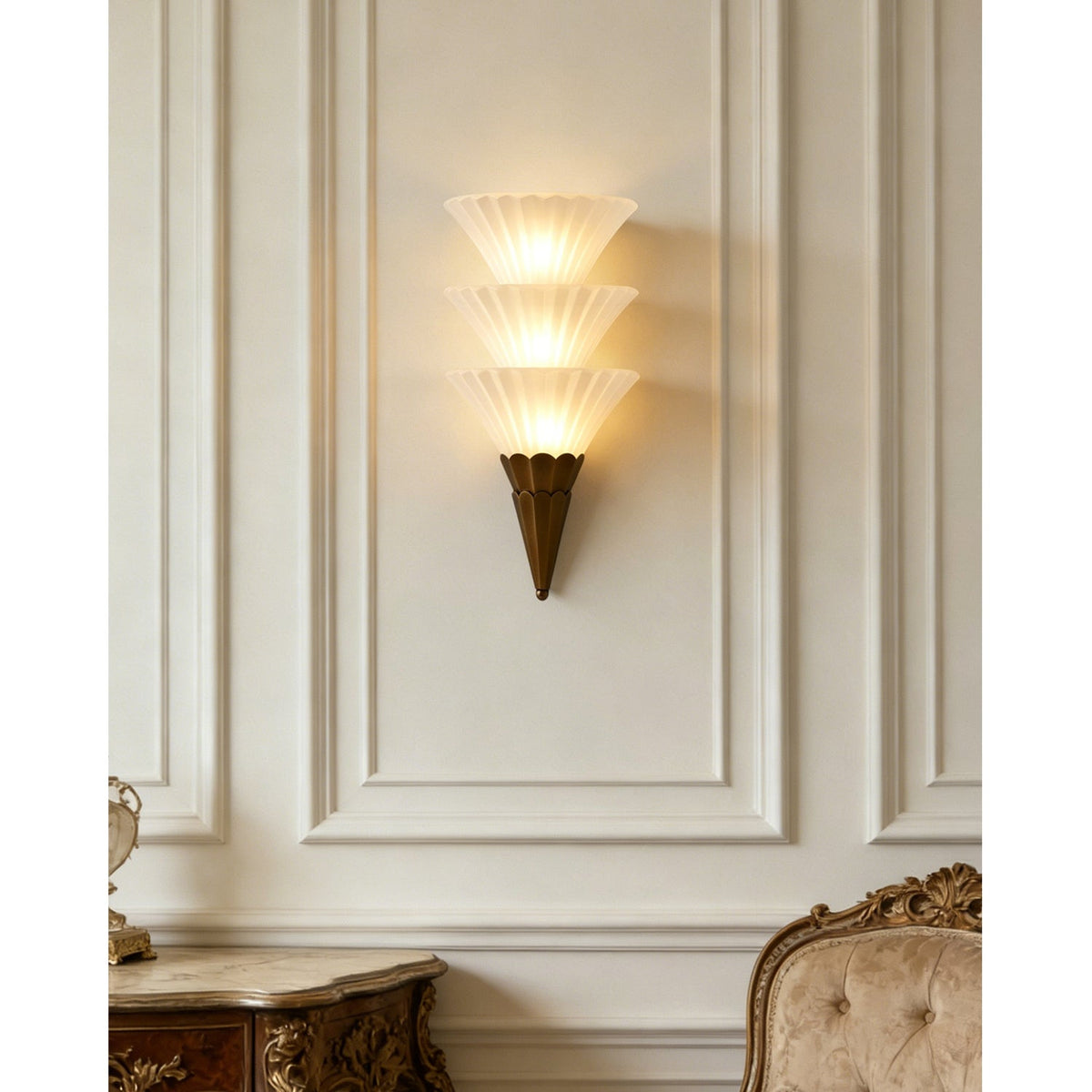 Lit Clérachic wall sconce with three stacked glass cones on a panelled cream wall, above a carved console and chair.