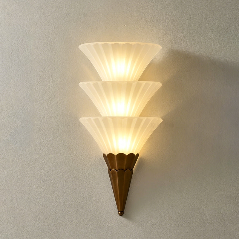 Three-cone Clérachic wall sconce lit warm, the stacked ribbed glass casting a soft shadow on a grey wall.