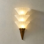 Three-cone Clérachic wall sconce lit warm, the stacked ribbed glass casting a soft shadow on a grey wall.