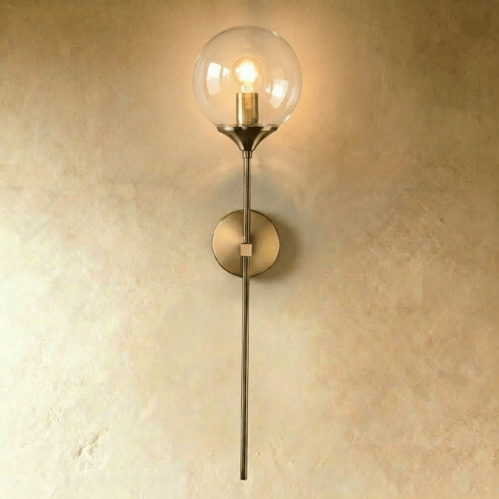 Clérachic wall sconce in brushed brass with a clear glass globe shade, straight stem lit warm white against a plaster wall.