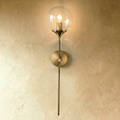 Clérachic wall sconce in brushed brass with a clear glass globe shade, straight stem lit warm white against a plaster wall.