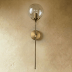 Clérachic wall sconce in brushed brass with a clear glass globe shade shown unlit, straight stem against a plaster wall.