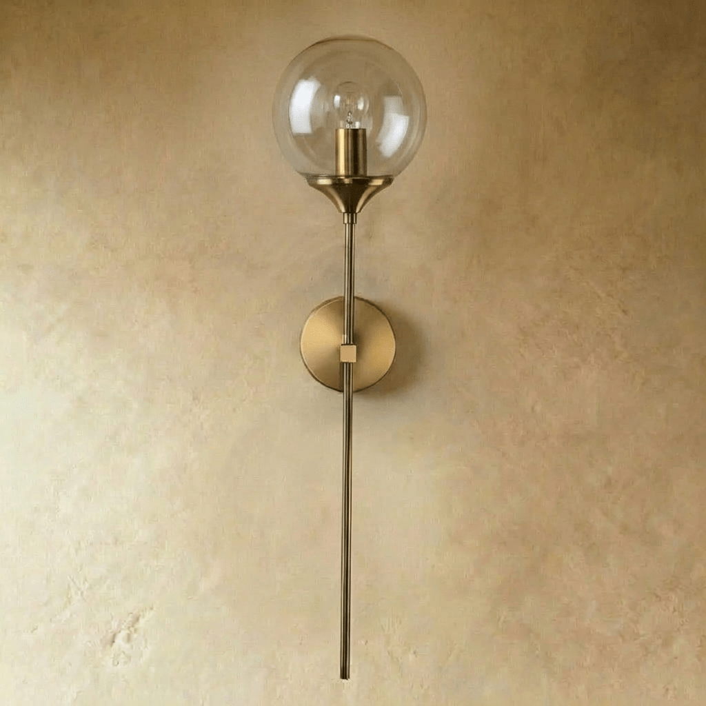 Clérachic wall sconce in brushed brass with a clear glass globe shade shown unlit, straight stem against a plaster wall.