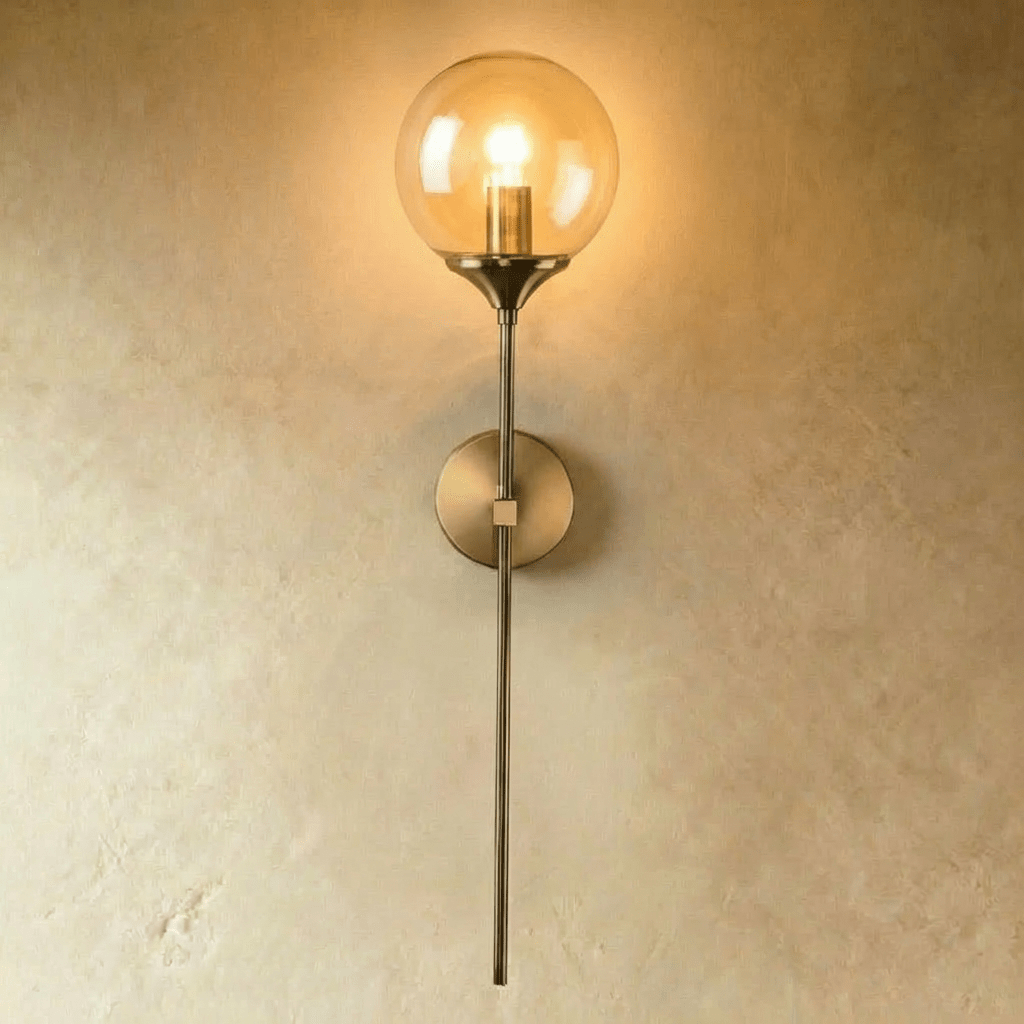 Clérachic wall sconce in brushed brass with a clear glass globe shade glowing warm white on a straight vertical stem.