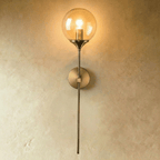 Clérachic wall sconce in brushed brass with a clear glass globe shade glowing warm white on a straight vertical stem.
