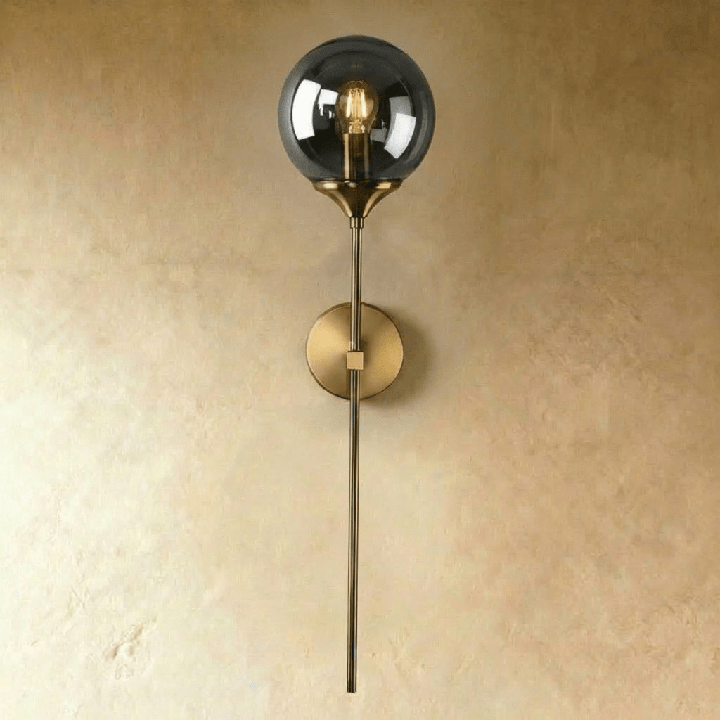 Clérachic wall sconce in antique brass with a smoked gray glass globe shade lit warm white on a straight stem.