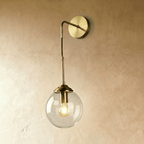 Clérachic wall sconce in brass with a clear glass globe shade on a bent drop arm, shown unlit against a plaster wall.