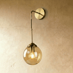 Clérachic wall sconce in brass with an amber glass globe shade on a bent drop arm, glowing warm white on a plaster wall.