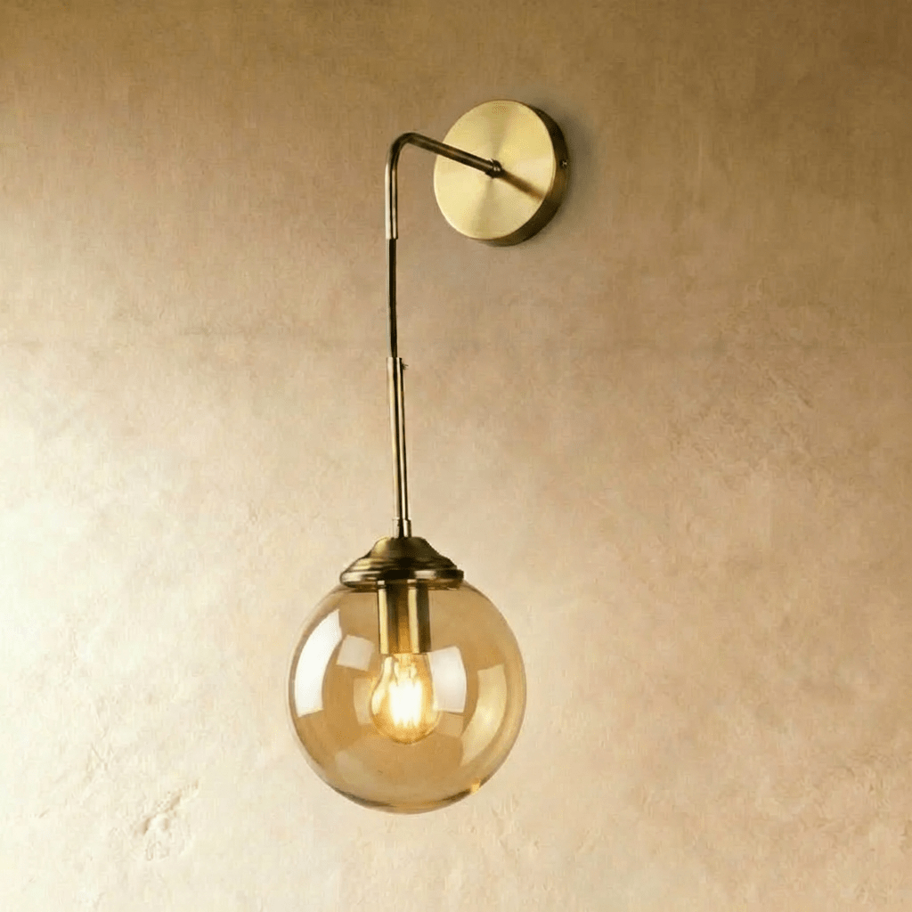 Clérachic wall sconce in brass with an amber glass globe shade on a bent drop arm, glowing warm white on a plaster wall.