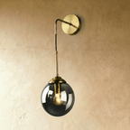 Clérachic wall sconce in brass with a smoked gray glass globe shade on a bent drop arm, lit warm white on a plaster wall.