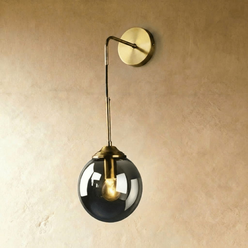 Clérachic wall sconce in brass with a smoked gray glass globe shade on a bent drop arm, lit warm white on a plaster wall.