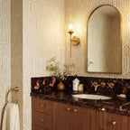 Clérachic wall sconce in brass with a clear glass globe shade lit warm white beside a bathroom vanity mirror and patterned wallpaper.
