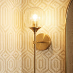 Clérachic wall sconce in brass with a clear glass globe shade lit warm white, seen close beside a mirror on patterned wallpaper.
