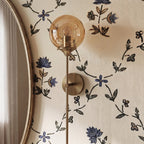 Clérachic wall sconce in brass with an amber glass globe shade lit warm white, seen close beside an oval mirror on floral wallpaper.