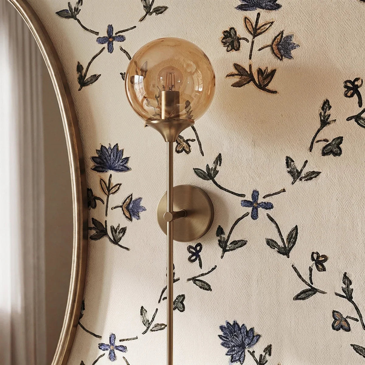 Clérachic wall sconce in brass with an amber glass globe shade lit warm white, seen close beside an oval mirror on floral wallpaper.