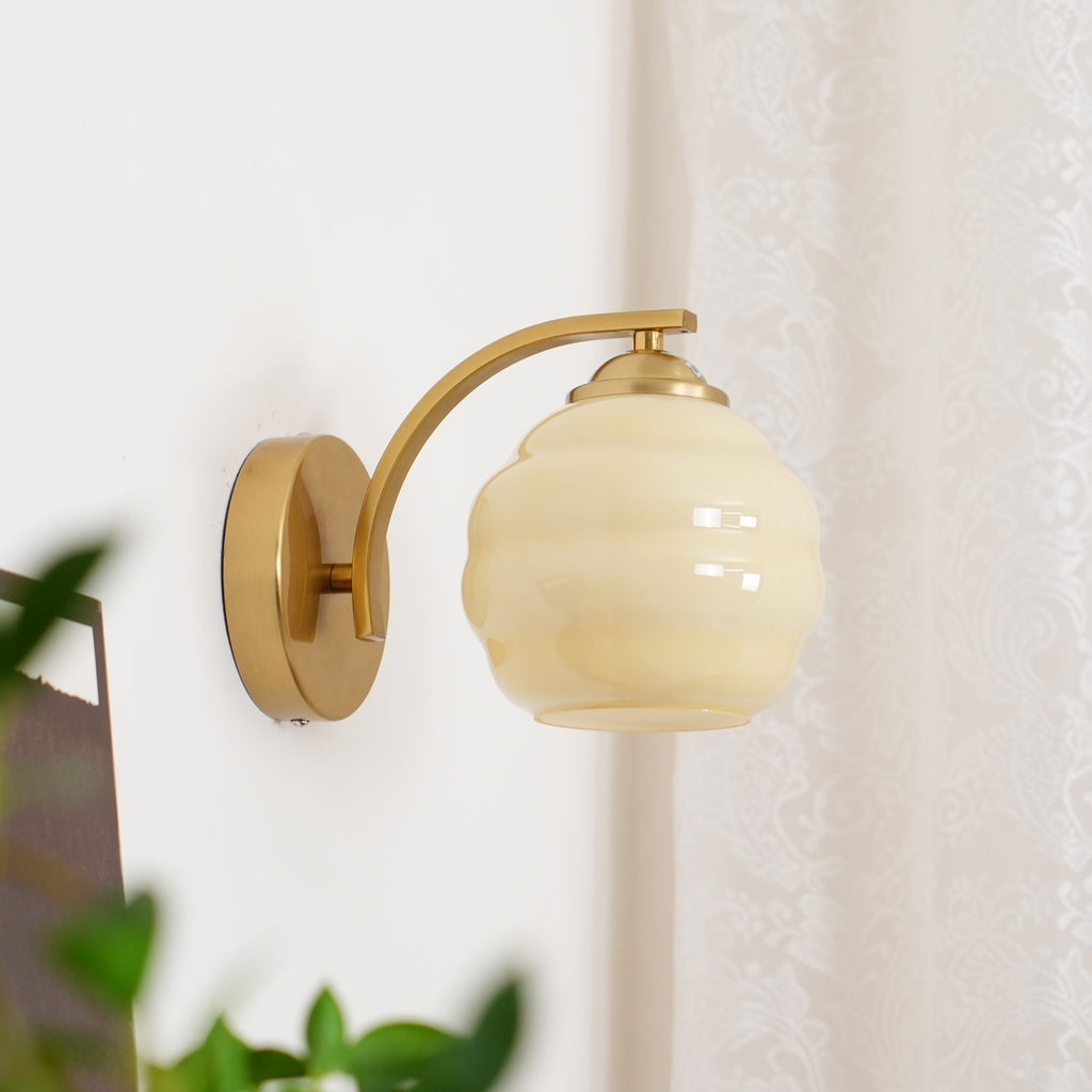 Unlit Clérachic gold wall sconce with a beige lantern shade beside a paisley curtain and green leaves.