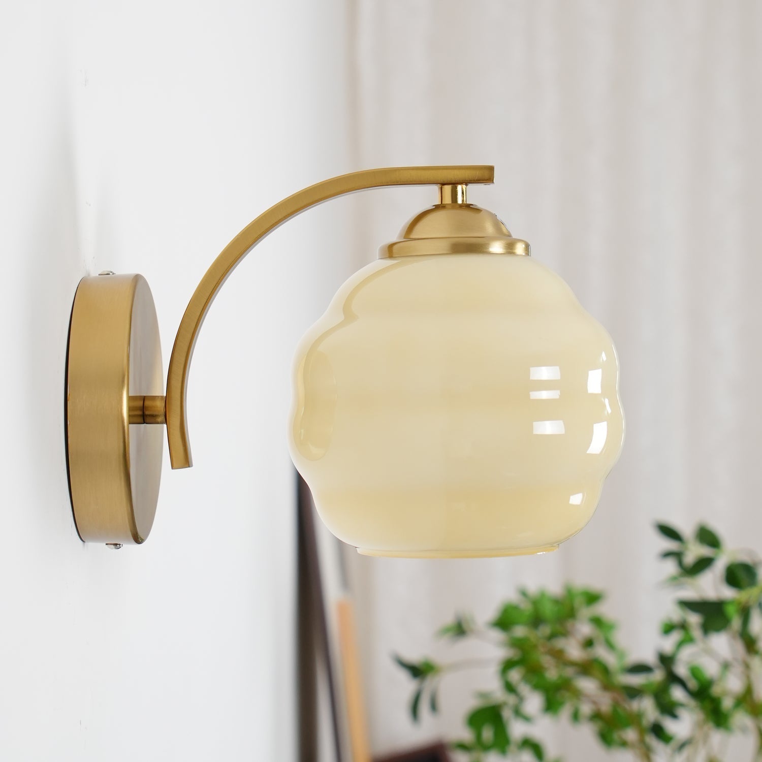 High-angle view of the Clérachic gold wall sconce and beige lantern shade, a plant and curtain behind.