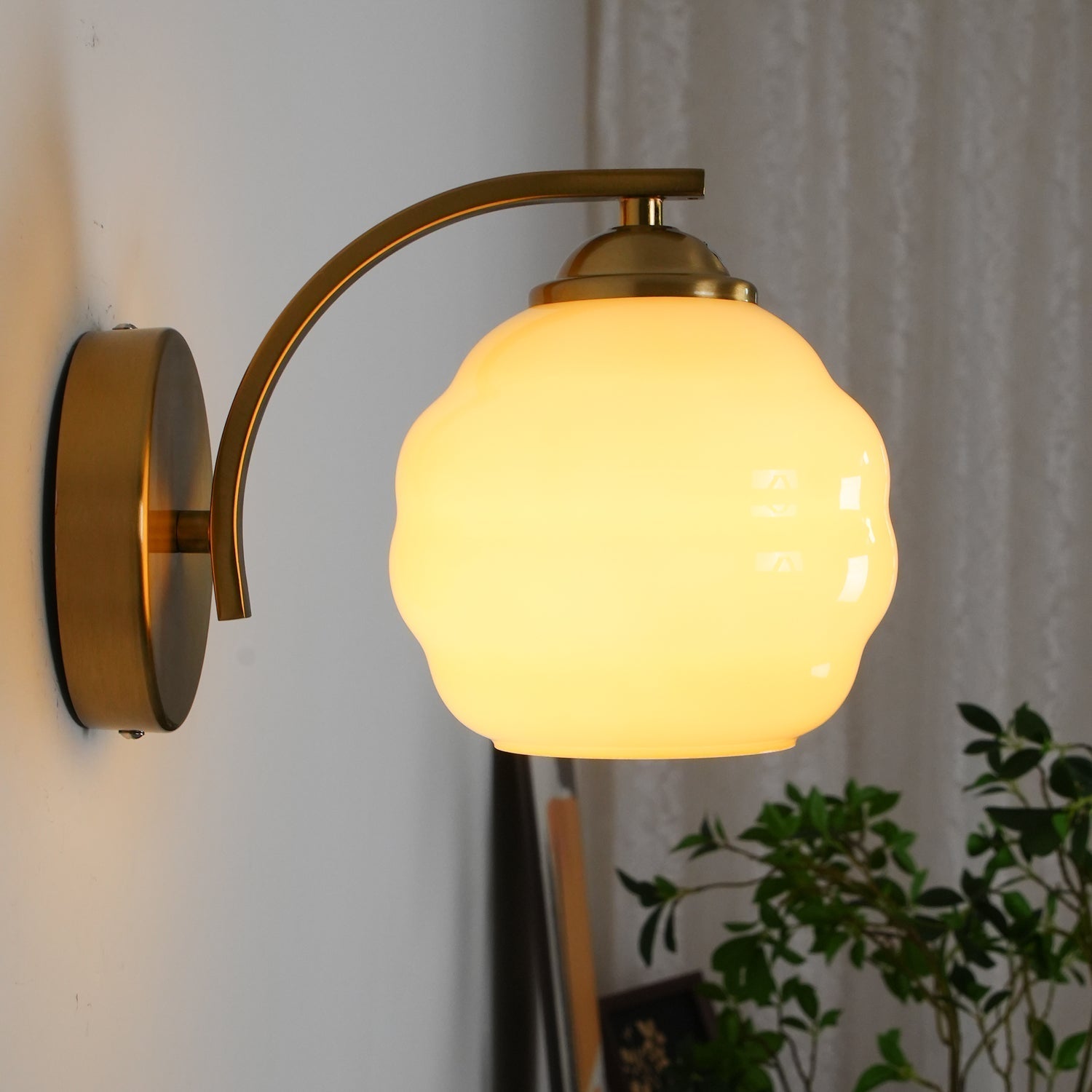 Lit Clérachic gold wall sconce with a creamy beige lantern shade beside a white curtain and houseplant.