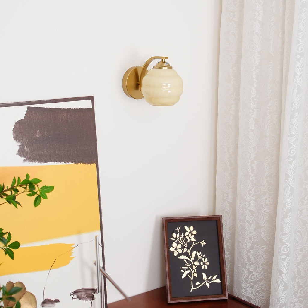 Clérachic gold wall sconce with a beige lantern shade above a floral print on dark wood, a lace curtain at right.
