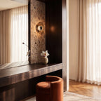Clérachic round glass wall light glowing on floral wallpaper above a dark wood dressing table with a velvet stool.