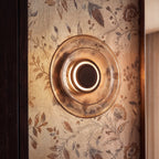 Clérachic round layered glass wall light close up, a lit ring around its dark centre on botanical wallpaper.