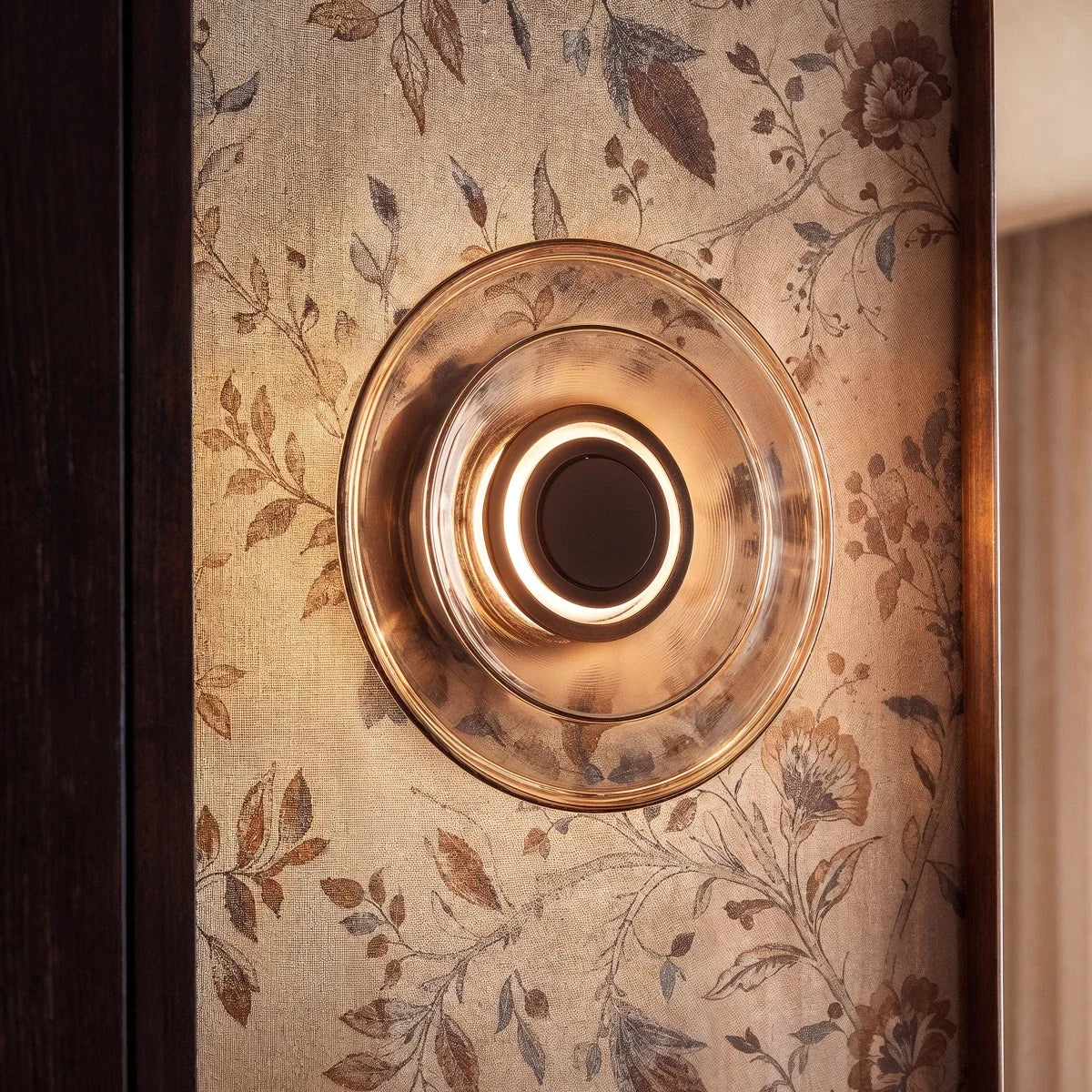 Clérachic round layered glass wall light close up, a lit ring around its dark centre on botanical wallpaper.