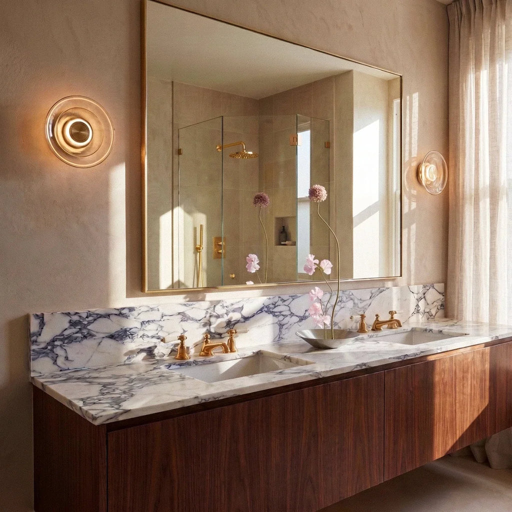 Clérachic pair of round amber glass wall lights flanking a large gold-framed mirror above a marble-topped counter.