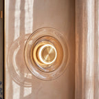 Clérachic round cognac glass wall light close up, a glowing ring around a brushed brass centre beside a linen curtain.