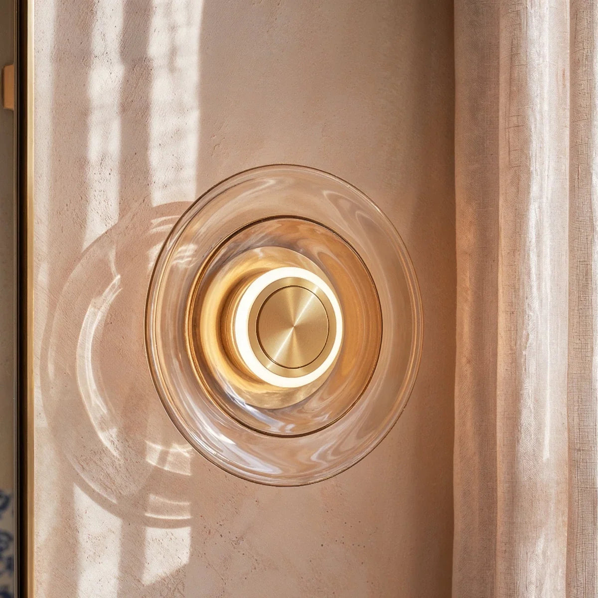 Clérachic round cognac glass wall light close up, a glowing ring around a brushed brass centre beside a linen curtain.