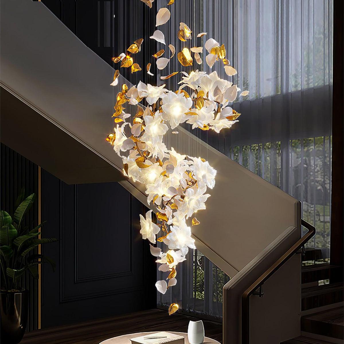 Beside a dark staircase and sheer curtains, this Clérachic chandelier hangs in a long spiral of lit white petals and brass leaves.