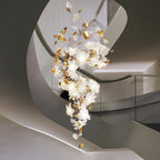 Above a sweeping pale curved staircase, a Clérachic chandelier drops in a slim cascade of glowing white petals and gold leaves.