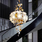 The rounded white petal cluster of this Clérachic chandelier glows over a black curved stair rail, brass leaves falling beneath it.