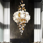 Against a dark stone wall between sheer curtains, a Clérachic chandelier shows a full round cluster of white petals above hanging leaves.
