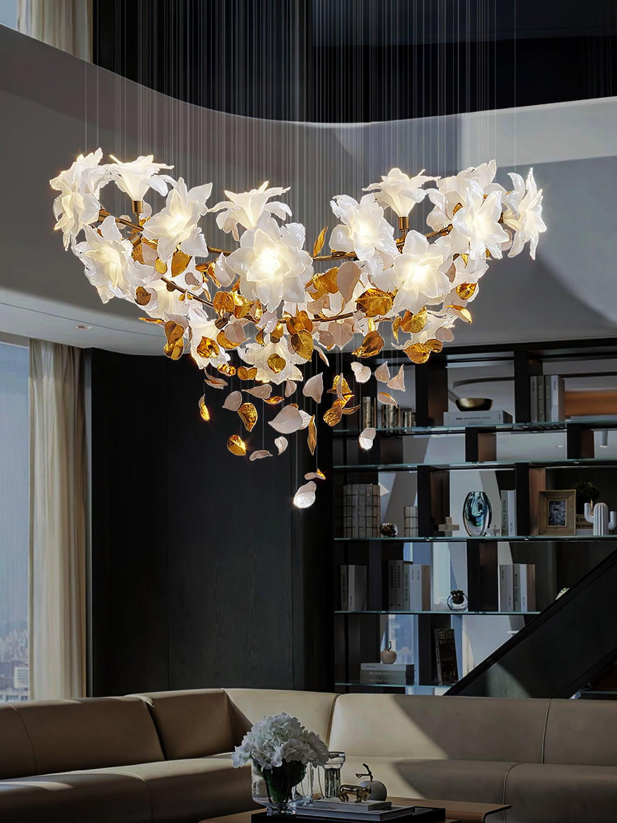 A wide, low Clérachic chandelier of white petals and brass leaves spreads over a living room with a brown sofa and open shelving.