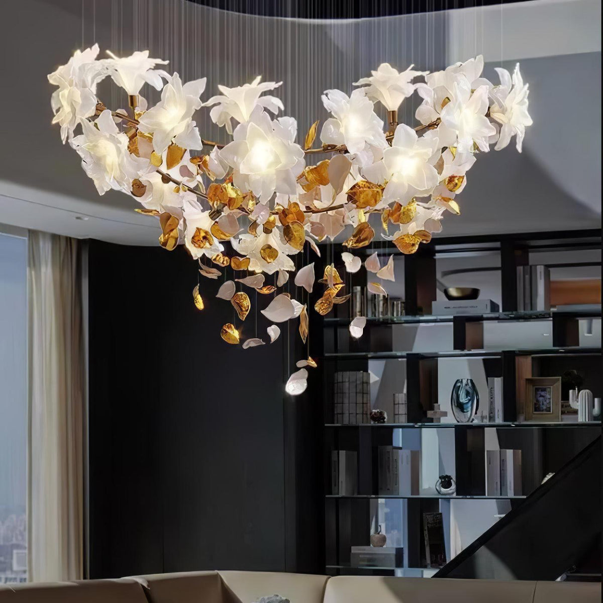 Close in on a wide, lit Clérachic chandelier whose white petal blooms sit on a brass ring above a dark bookshelf and sofa.