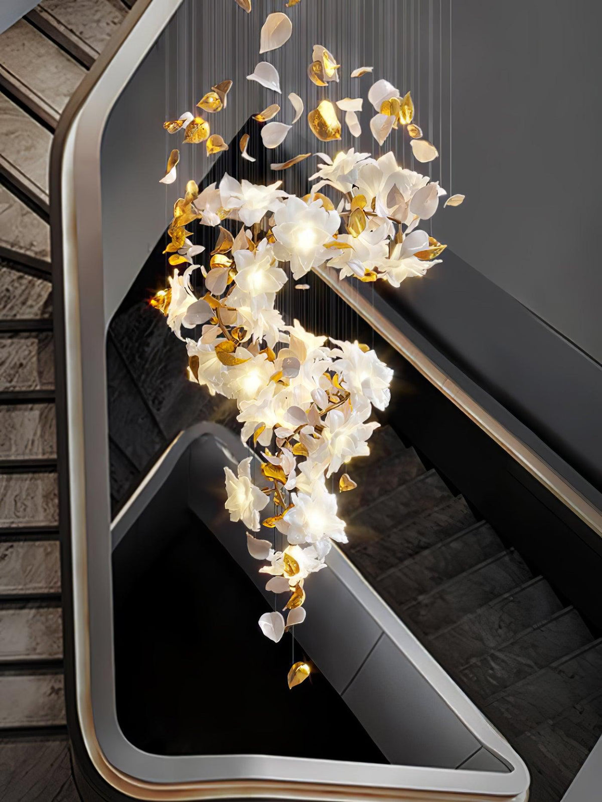 Seen from above a black staircase, this Clérachic chandelier curls in a lit S of white petals and gold leaves beside a pale handrail.