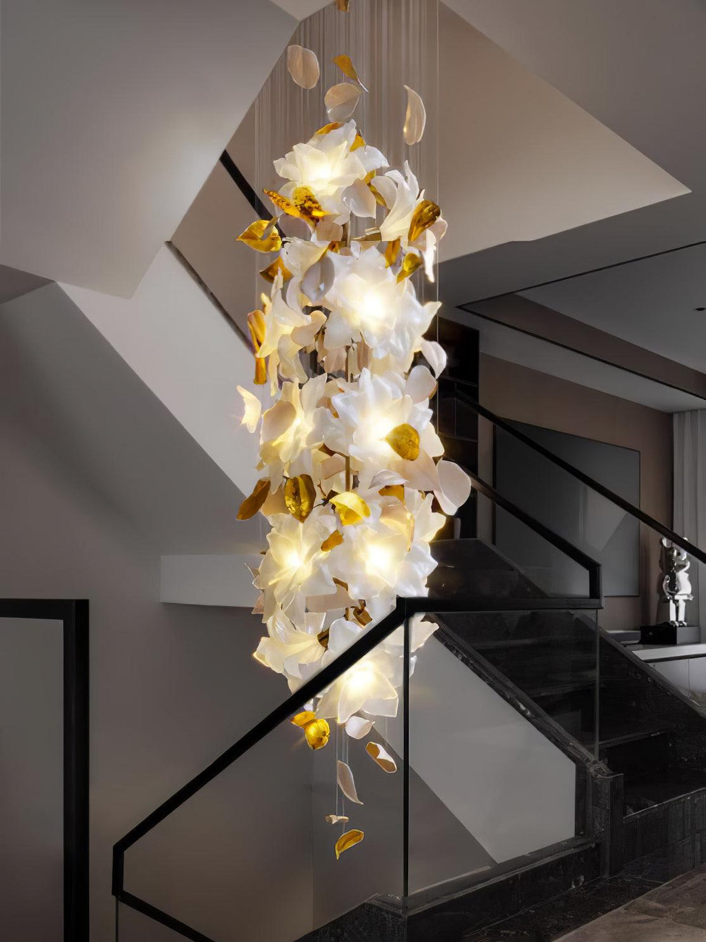 Tall and narrow, this Clérachic chandelier fills a stairwell with glowing white petals and brass leaves beside a glass balustrade.