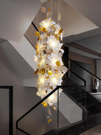 Tall and narrow, this Clérachic chandelier fills a stairwell with glowing white petals and brass leaves beside a glass balustrade.