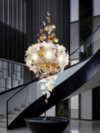 A rounded cluster of lit white petals with brass leaves below, this Clérachic chandelier hangs beside a black spiral staircase.
