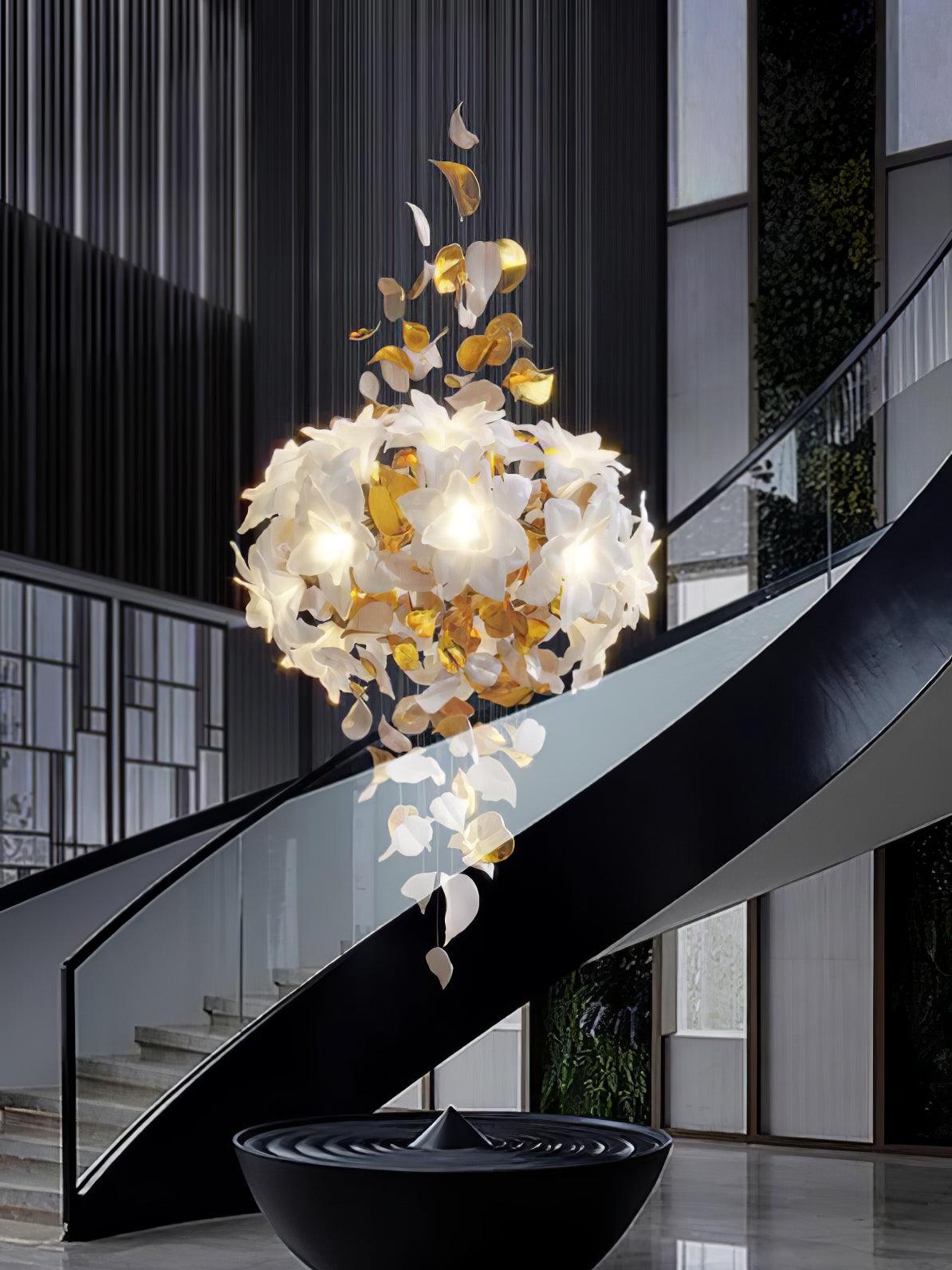 A rounded cluster of lit white petals with brass leaves below, this Clérachic chandelier hangs beside a black spiral staircase.